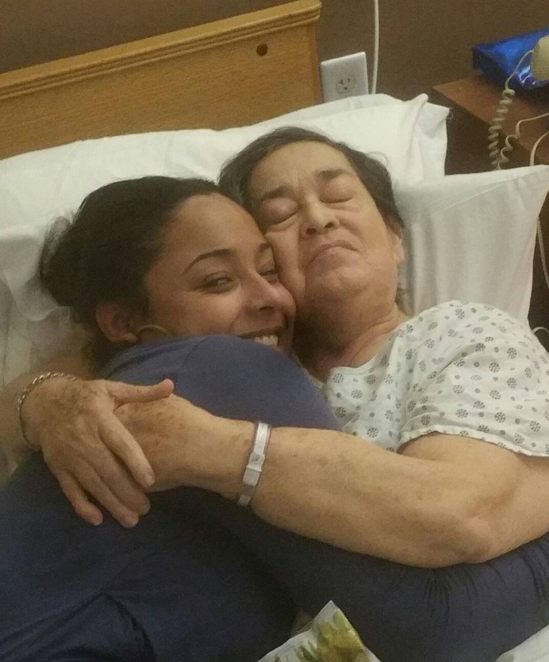 An image of Rebekah smiling wide and hugging her grandmother, her Tata for whom she does caregiving, who is in a hospital gown in bed.