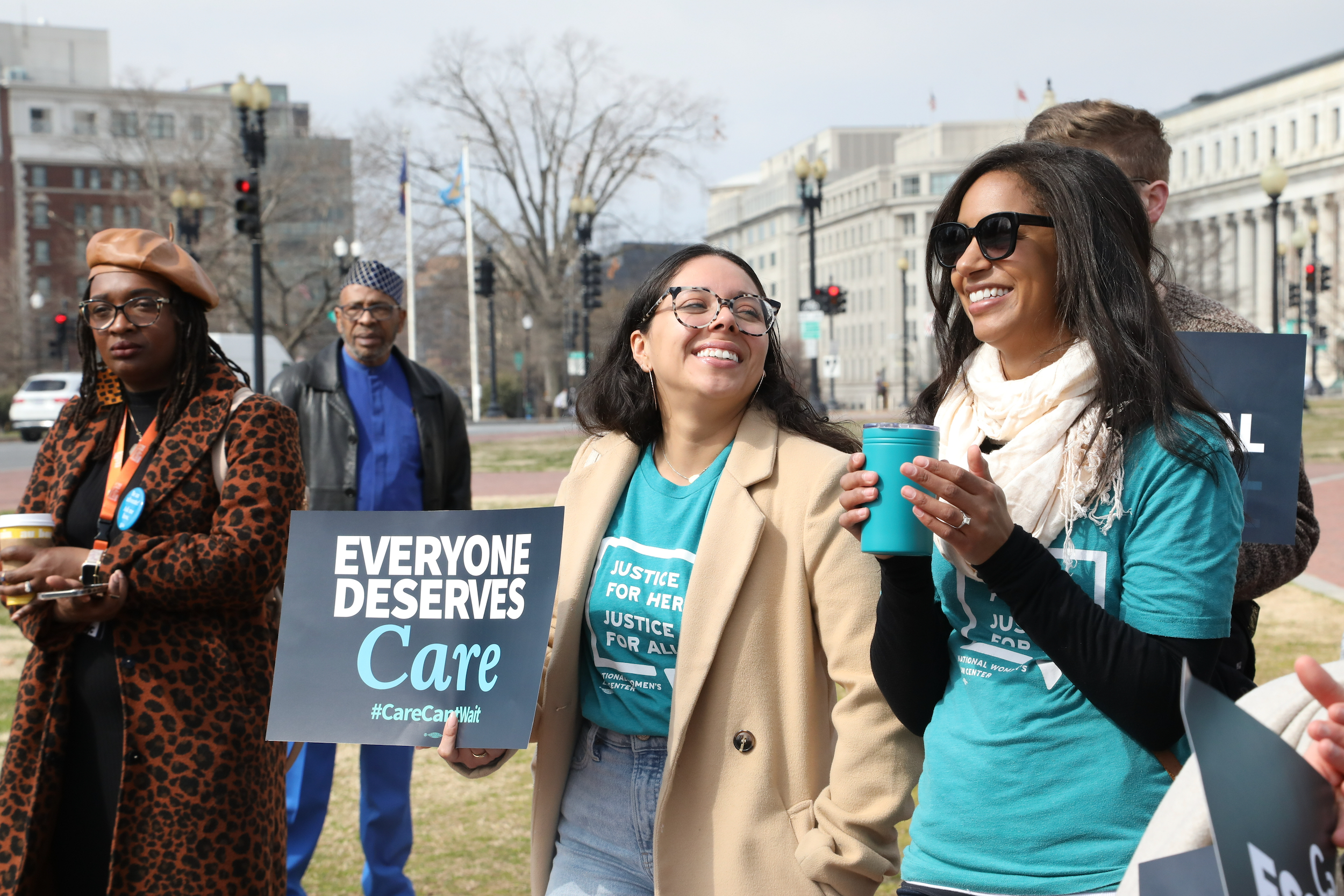 Two women smile together as one holds sign saying "Everyone deserves care"